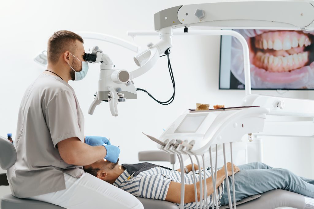 young-man-with-patient-bib-dental-chair-dentist-who-sits-him-he-looks-his-teeth-using-dental-microscope-holds-dental-bur-mirror